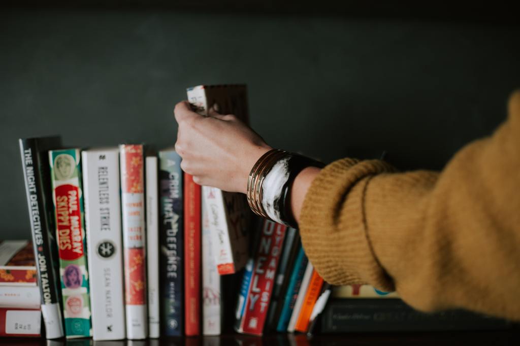 A person reaching for a book on a shelf filled with various titles and colorful book spines.