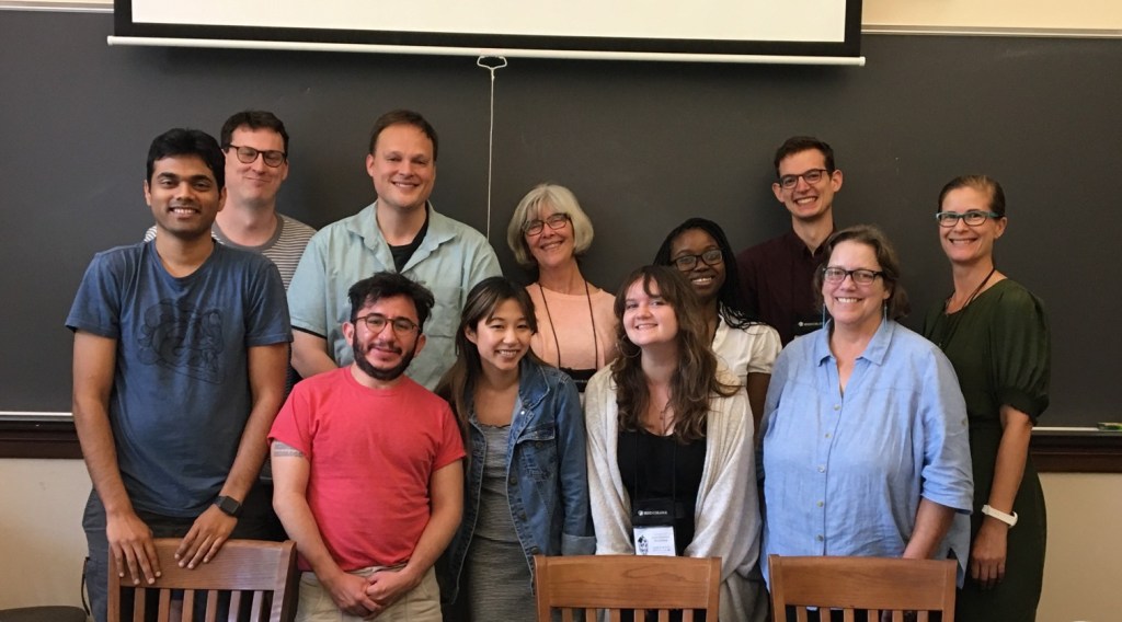 Group photo of participants at a writing workshop, standing and sitting in front of a chalkboard.